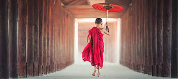 Asienwissenschaften Das Bild zeigt einen asiatischen Jungen durch den Flur eines Gebäudes gehen. Fotografiert ist die Rückenansicht des Jungen. Er trägt pinkfarbene Mönchskleidung, ist barfuß und hält einen roten Schirm. Der Flur besteht aus hölzernen Balken mit Zwischenräumen, durch die Licht fällt.