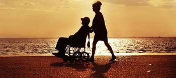 Bachelor Sonderpädagogik Bild von einem Strand im Sonnenuntergang. Im Zentrum des Bildes die Konturen zweier Personen. Die eine Person schiebt die andere in einem Rollstuhl an dem Strand entlang.